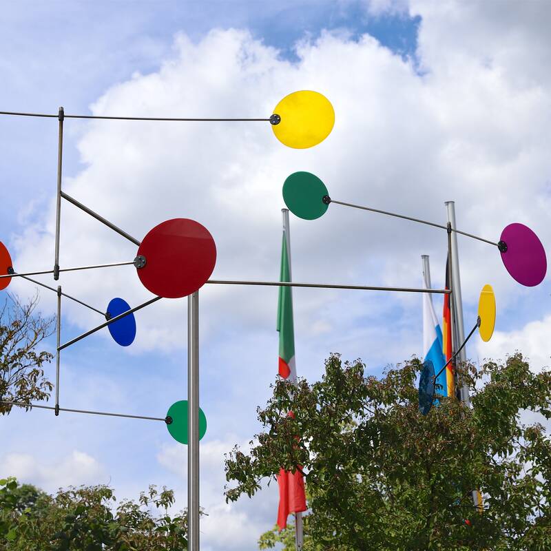 Eine farbenfrohe Skulptur aus sich drehenden, kreisförmigen Elementen in verschiedenen Farben steht vor einem bewölkten Himmel und ist von Bäumen umgeben.