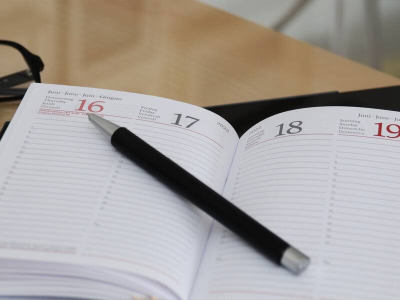 Ein geöffneter Terminplaner zeigt die Daten 16. bis 19. Juni 2022, mit einem schwarzen Stift darauf liegend und einer Brille im Hintergrund.