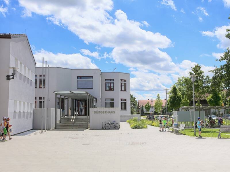 Das Bild zeigt das moderne Gebäude eines Bürgerhauses mit einer offenen Platzanlage und mehreren Menschen bei sonnigem Wetter.