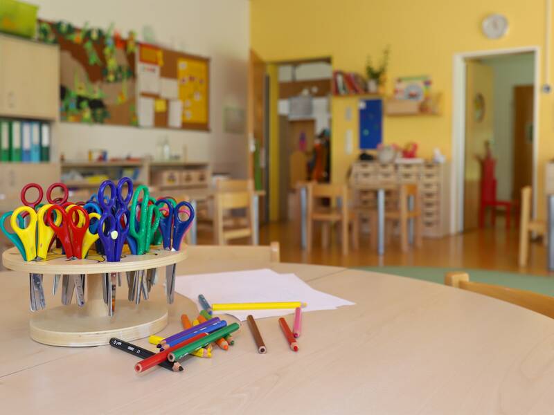 Ein buntes Sortiment an Scheren und Stiften liegt auf einem Tisch in einem hellen Klassenzimmer mit gelben Wänden.