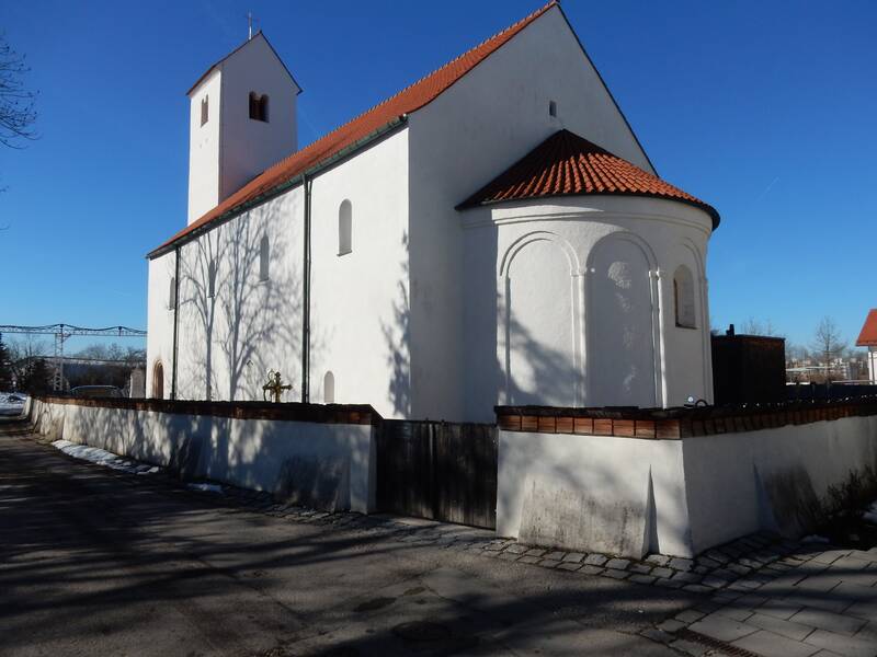 Ein weißes Kirchgebäude mit rotem Ziegeldach und Glockenturm steht unter klarem blauen Himmel, umgeben von einer niedrigen Mauer.
