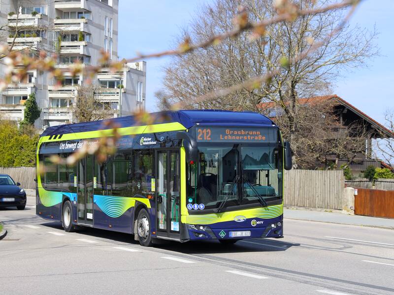 Ein blauer Linienbus mit der Aufschrift „Grasbrunn Lehnerstraße“ fährt auf einer Straße an einem Wohngebiet entlang, während im Hintergrund ein Auto zu sehen ist.