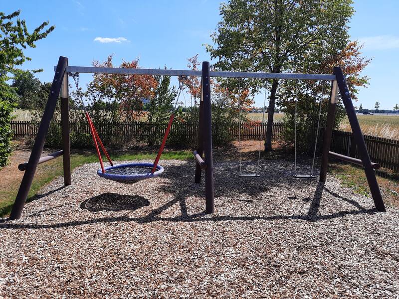 Ein Spielplatz mit einer Doppelschaukel, bestehend aus einem Nest und einem traditionellen Sitz, steht auf einem mit Holzhackschnitzeln bedeckten Boden vor einem Hintergrund aus Bäumen und einem Holzzaun.