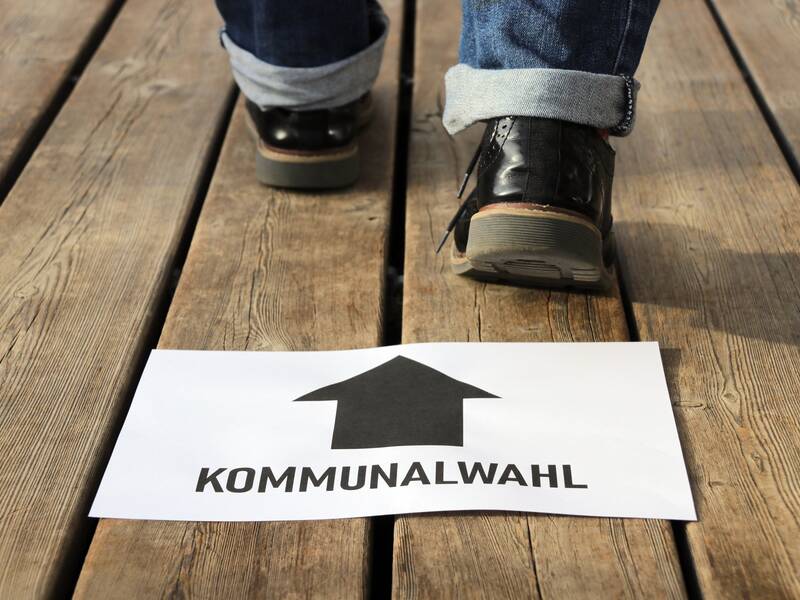 Ein Paar Beine in Jeans und Schuhen geht einen Holzsteg entlang, wobei ein Schild mit einem Pfeil und dem Text "KOMMUNALWAHL" auf dem Boden liegt.