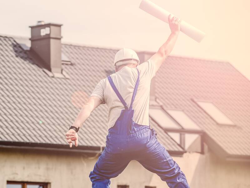 Ein Bauarbeiter in blauer Latzhose springt vor einem neu gebauten Haus und hält eine gerollte Papierrolle in die Luft.